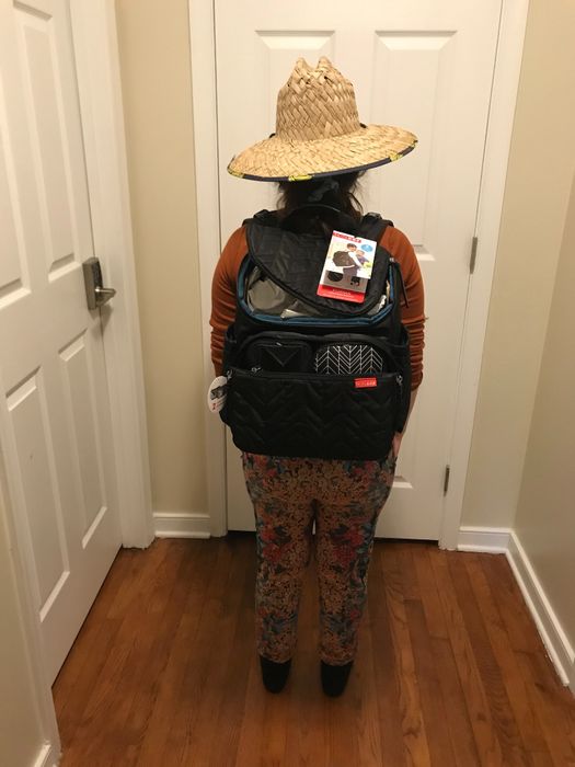 A person stands indoors with their back to the viewer, wearing a large backpack adorned with tags and a wide-brimmed hat. Their patterned pants add a bold touch to the outfit.