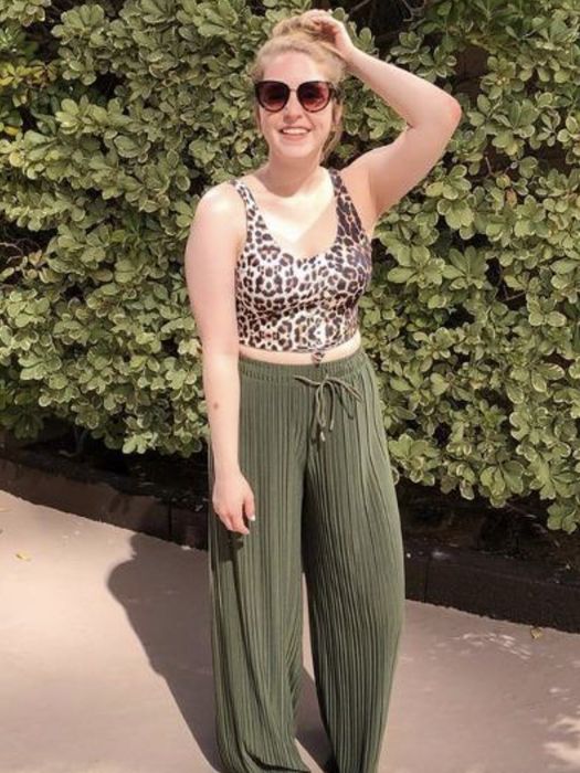 A woman in a leopard print top and green pleated pants strikes a pose in front of a leafy backdrop, hand placed on her head.