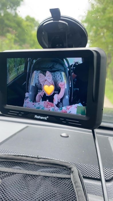 A baby in a car seat, visible through a baby monitor screen mounted on the dashboard, with a heart emoji playfully covering their face.