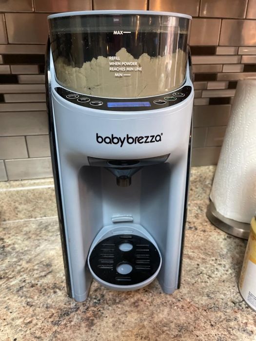 The Baby Brezza Formula Pro sits on a kitchen countertop, with a paper towel roll in the background, and automates the process of preparing baby formula.