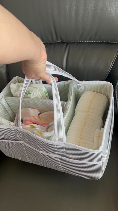 A hand gripping a striped diaper caddy, filled with diapers, packaged baby wipes, and neatly organized into various compartments for convenience.