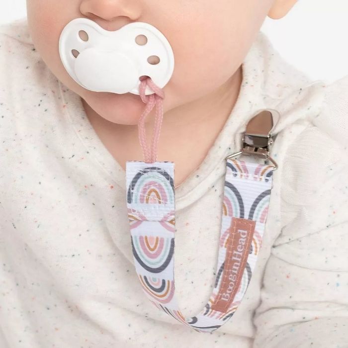 A pacifier clip with a rainbow design attached to a pacifier in a baby’s mouth, with the clip resting on their shirt