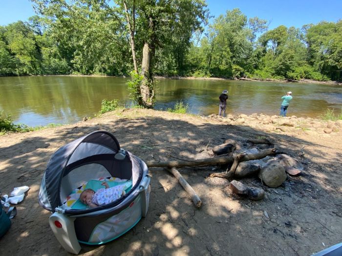 A baby peacefully sleeps in a portable crib set up by a riverside campsite, with two people fishing in the background, surrounded by trees.