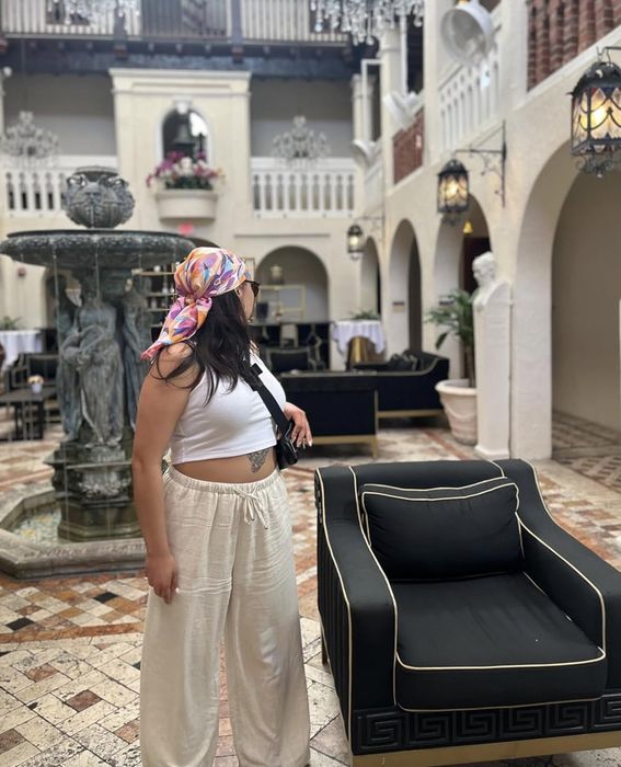 A woman in a headscarf and casual outfit stands in a courtyard with a fountain and balcony in the background.