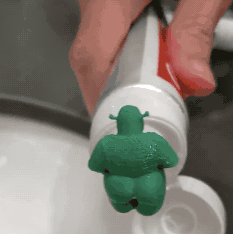 GIF of a limbless plastic Shrek figure on a toothpaste tube, with toothpaste squeezing out of its backside onto a toothbrush