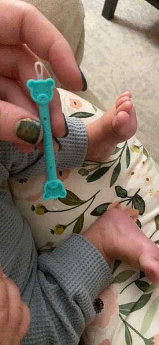 A hand holding a blue baby teething toy shaped like a bear, placed near a baby's feet. The baby is dressed in a waffle-textured outfit.