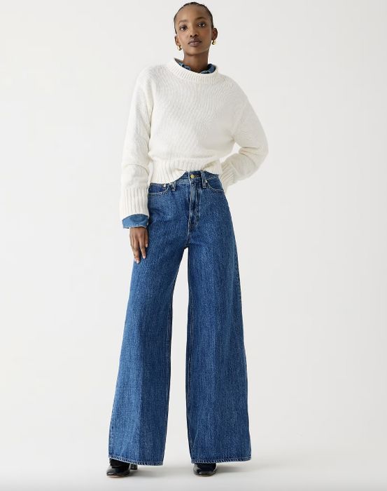 A woman is standing in a white sweater and wide-leg jeans, hands in her pockets, wearing black footwear.