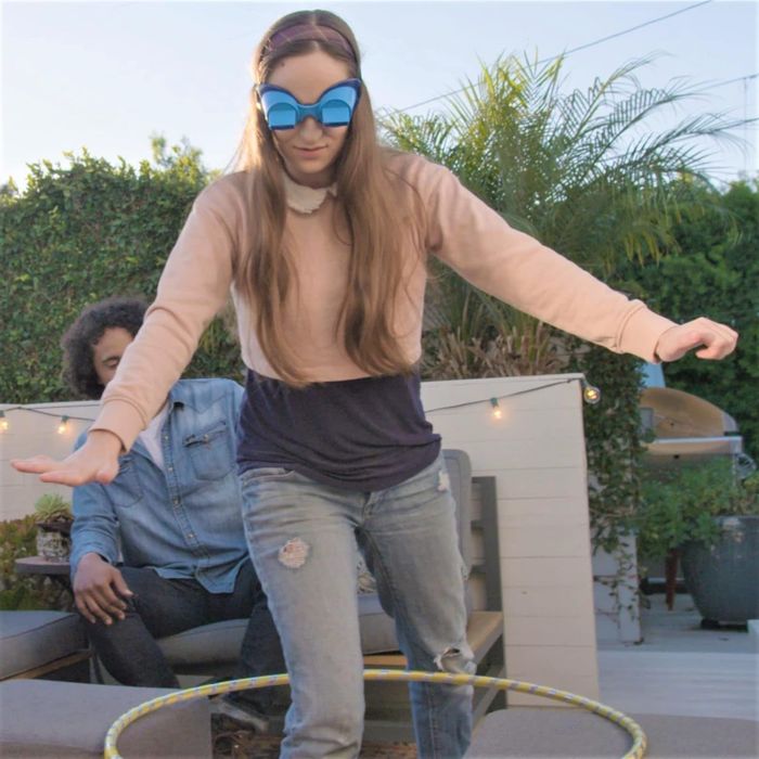 A woman is hula hooping in casual clothes while a man lounges in the background. They are enjoying a bright, sunny day outdoors.