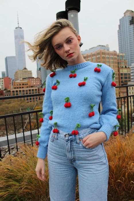 A model wearing a light blue sweater decorated with 3D knit cherries all over it.