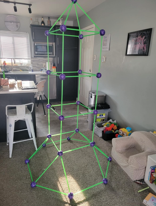 A large green and purple geometric play structure sits in a living room, surrounded by toys and household items, with the kitchen area visible in the background
