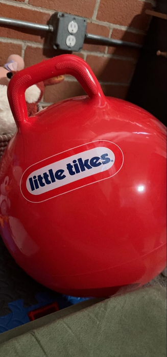 A red Little Tikes bouncing ball with a handle, resting against a brick wall backdrop.