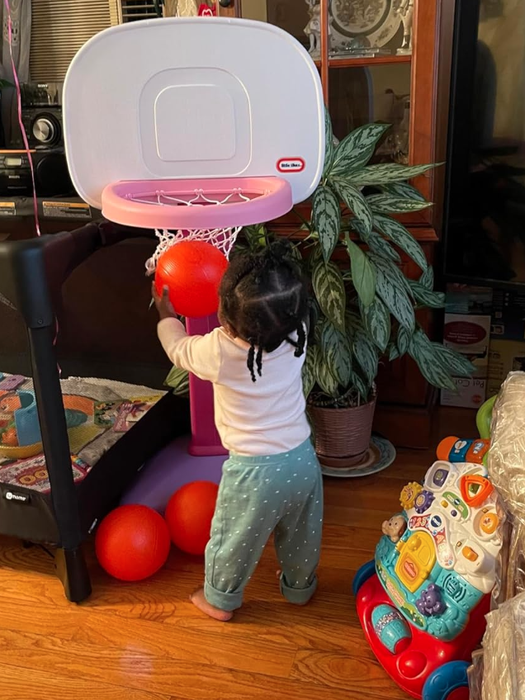 A young child in polka-dot pants is enjoying a mini indoor basketball hoop, happily shooting a red ball through the hoop. Nearby, there's a colorful baby activity walker.