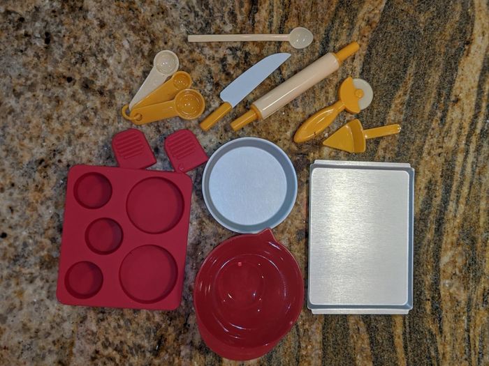 An assortment of miniature baking tools, including a silicone cupcake mold, measuring spoons, a metal tray, bowls, and a rolling pin, neatly arranged on a granite countertop.
