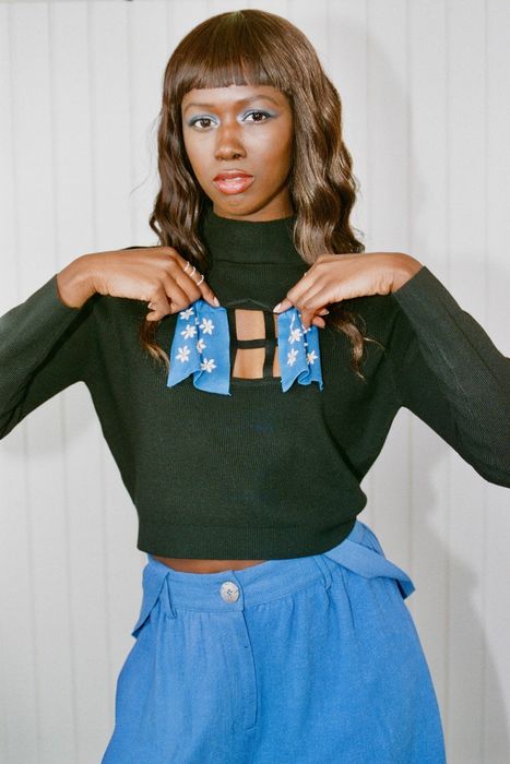 A model wearing a black cropped sweater featuring a window-shaped chest cutout with movable blue knit curtains.