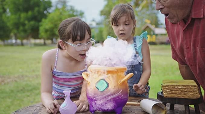 Two children and an adult watching a toy cauldron with smoke billowing out of it