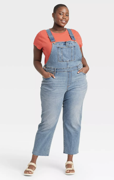 A model styled in denim overalls with a bright orange shirt underneath.