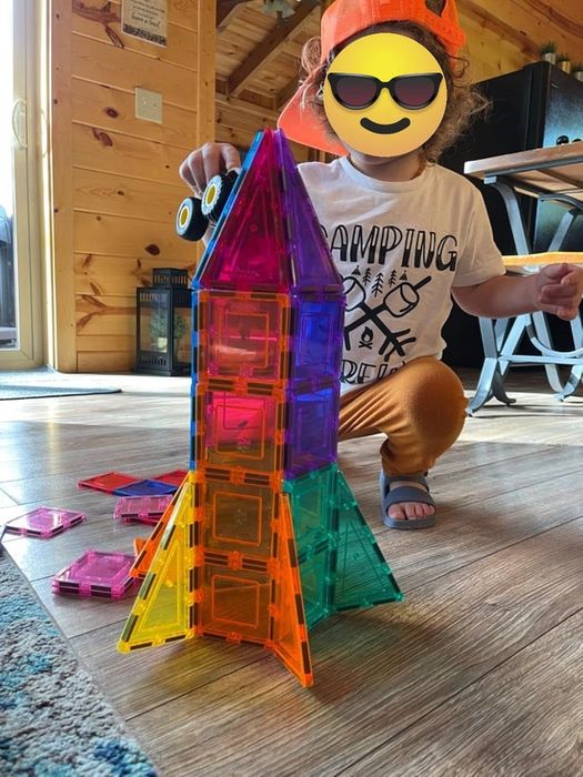 A child is constructing a vibrant magnetic tile rocket on a wooden floor indoors.