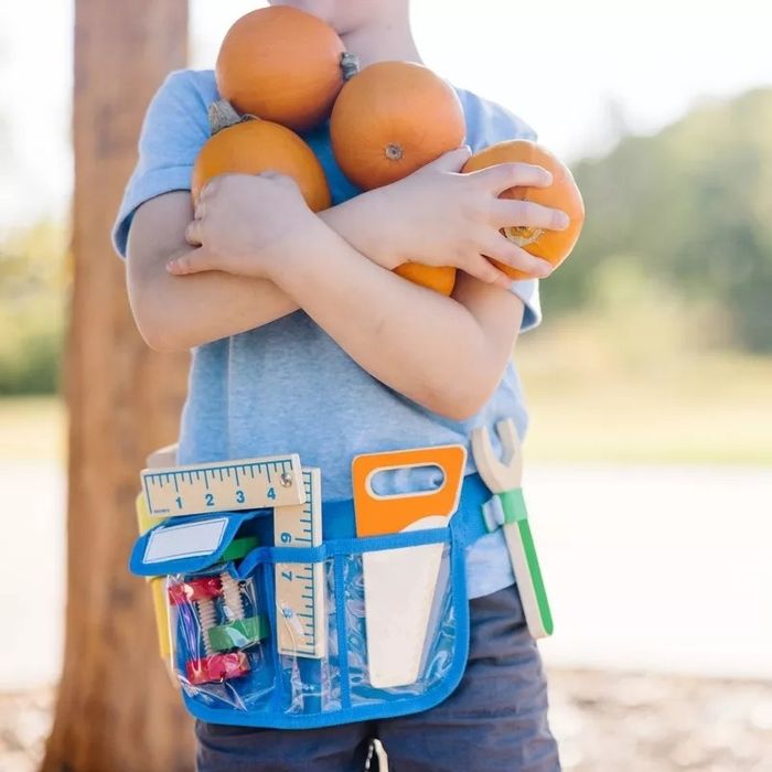 A child, dressed casually, holds three pumpkins and sports a blue tool belt filled with school supplies, set against an outdoor backdrop.