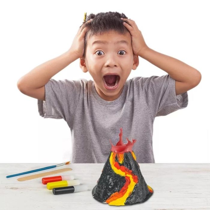 A boy in a gray t-shirt joyfully holds his head in excitement next to a toy volcano kit on a table, complete with brushes and various colored paints