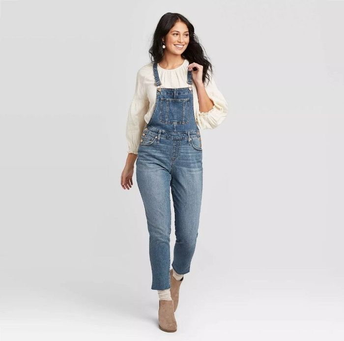A model showcasing a denim overalls paired with a flowing white long-sleeve shirt.
