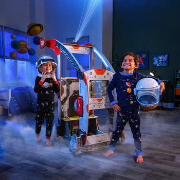 Two children wearing space helmets and pajamas, standing in front of the wooden spaceship set
