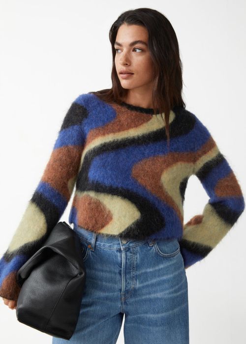 A model wearing a crewneck sweater with wide sleeves in a swirl pattern of cream, light brown, black, and blue.