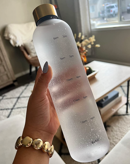 A reviewer holding a clear frosted white water bottle with subtle black measurement markings on the side and a gold twist-off lid.