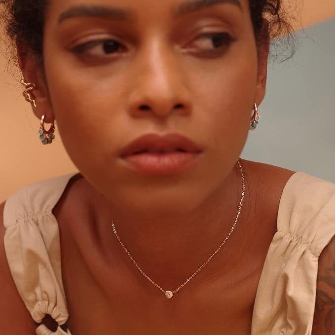 A model wearing a thin gold chain with a delicate heart pendant featuring the letter 'A' engraved inside.