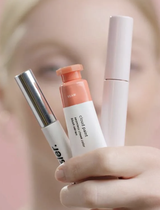 A model showcasing the peach Cloud Paint, Boy Brow, and Lash Slick mascara products from Glossier.