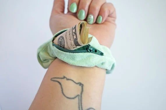 A person shows off their wrist with a green 'stashie' scrunchie, revealing cash tucked inside the zippered pocket.