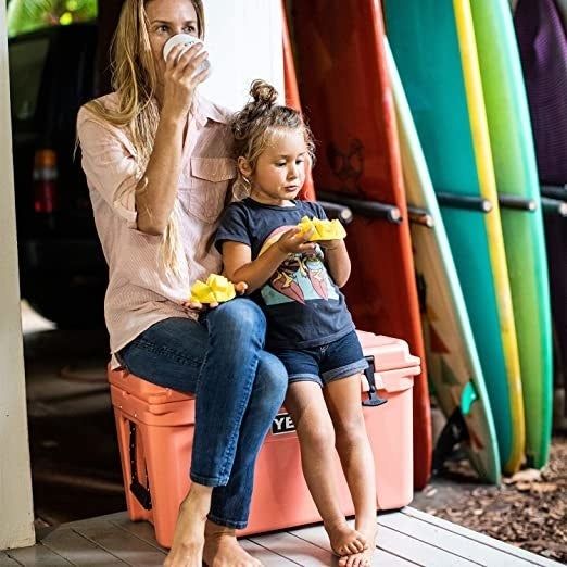 A woman and child sit together on a pink cooler, likely enjoying a relaxing break during an outdoor activity. The cooler serves as both a convenient place to store drinks and a spot to take a seat, adding comfort and practicality to their outing.