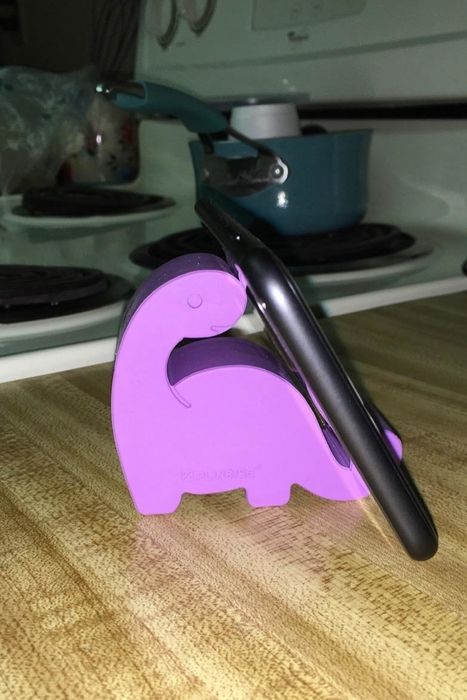 Reviewer's tiny purple dinosaur-shaped phone stand supporting a phone on a countertop