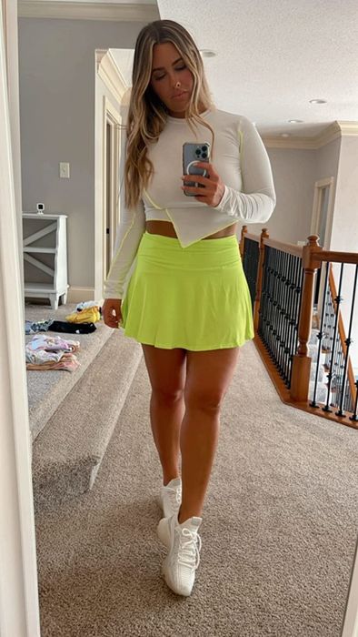 A reviewer is wearing a neon green tennis skirt, white sneakers, and a white long-sleeve crop top, standing by a wooden banister.