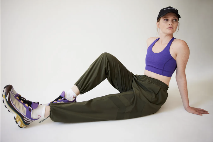 A model poses sitting with legs extended, showcasing green contrasting track pants, a bright purple cropped top, and black, yellow, and purple sneakers against a white backdrop.