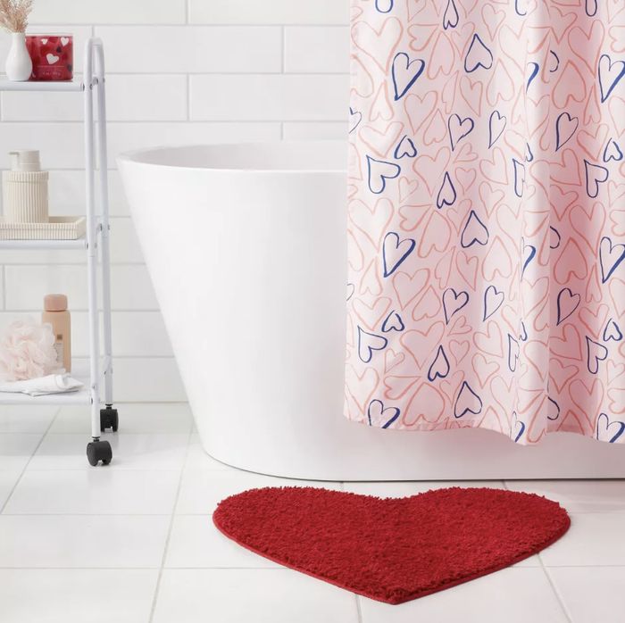 Bathroom with a white tub, heart-patterned shower curtain, metal shelf holding toiletries, and a red heart-shaped bath mat on the floor