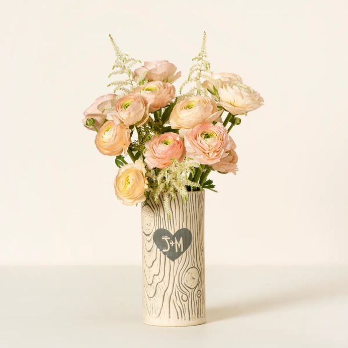 A beautiful bouquet of ranunculus and white flowers, arranged in a woodgrain-patterned vase engraved with a heart reading 'J+M.'