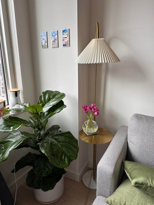 A stylish corner of a modern living room featuring a tall floor lamp, a potted plant, a small round gold table with pink tulips in a vase, and a section of a gray sofa.