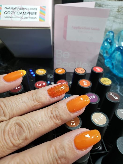 A reviewer's nails painted in a bright orange shade, with the kit and polish bottles in the background