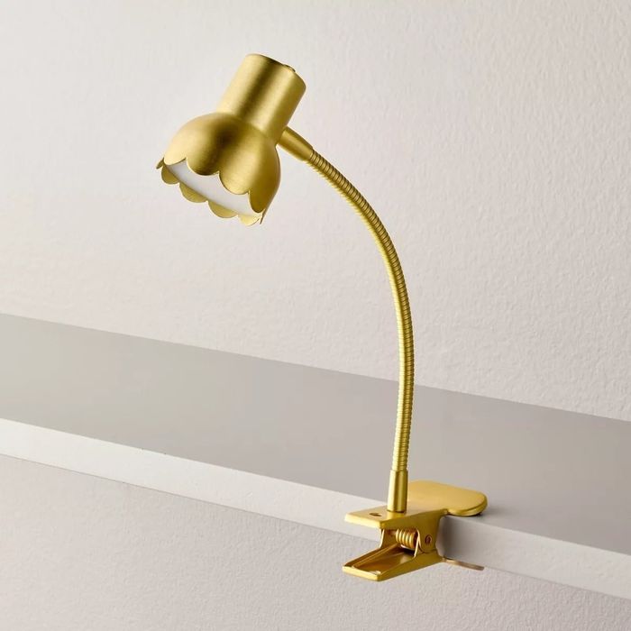 A gold-tone, scalloped-edge clip-on lamp attached to a shelf, featuring a flexible neck for adjustable lighting.