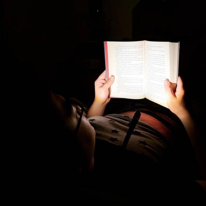 A reviewer lies in a completely dark room, with the neck reading light around their neck, casting light on the book in front of them.