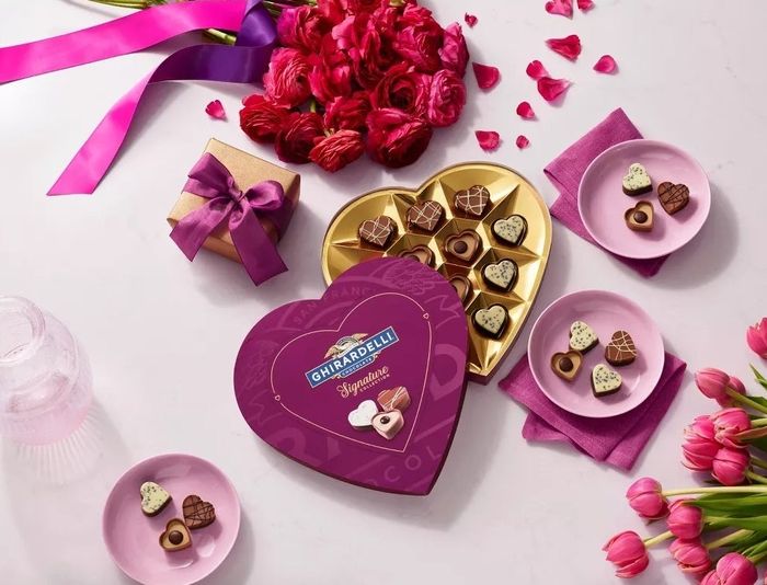 A heart-shaped Ghirardelli chocolate box, accompanied by flowers and a gift, set on a table — the perfect romantic gesture or Valentine’s Day gift.