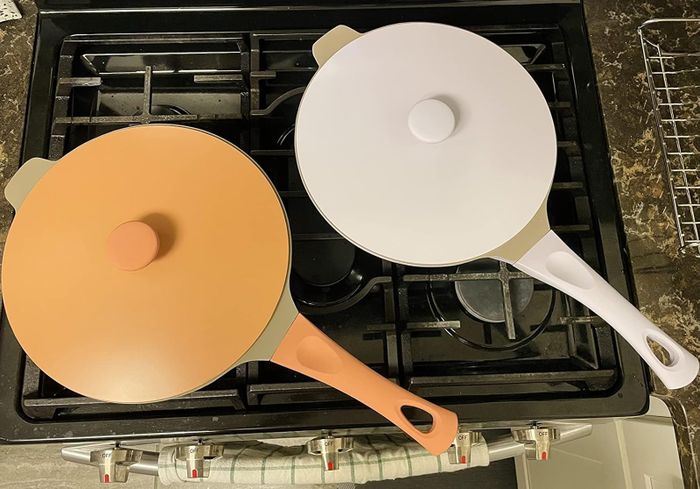 Two frying pans on a stovetop, one with an orange lid and the other with a white lid.