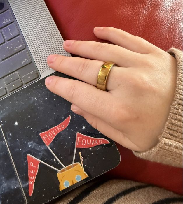 A hand adorned with a gold-tone Oura Ring Gen3 Horizon rests on a laptop keyboard, with a sticker reading 'Keep Moving Forward' surrounded by small flags.