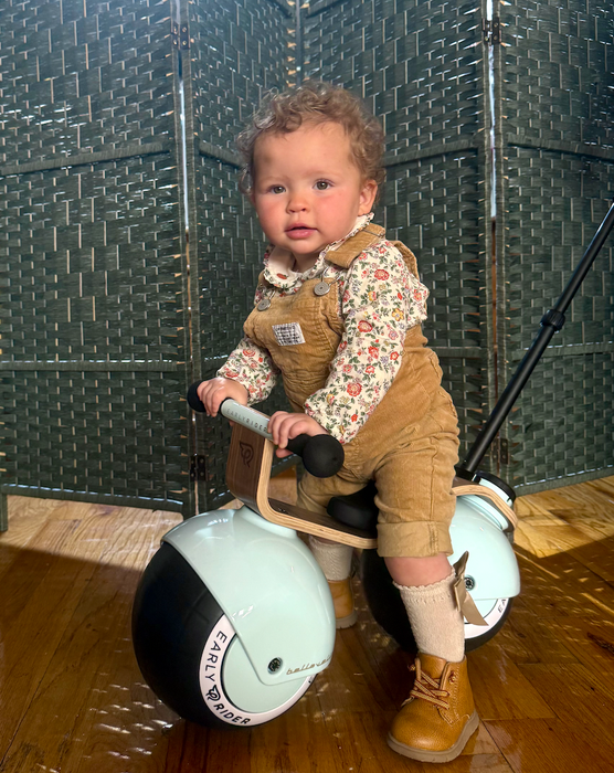 A toddler dressed in a floral shirt and corduroy overalls, sitting on a trendy ride-on toy indoors.