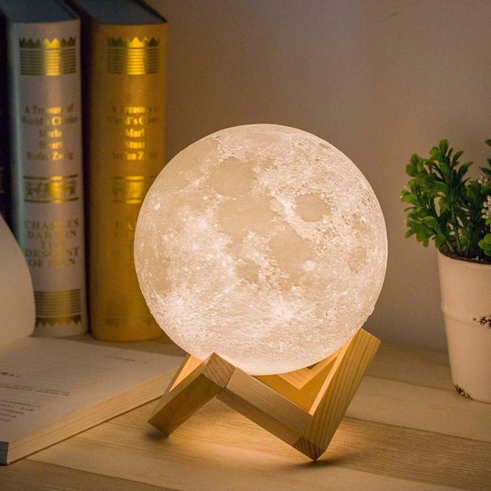 A moon lamp on a wooden base beside an open book and a small plant, creating a cozy ambiance for home decor and soft lighting