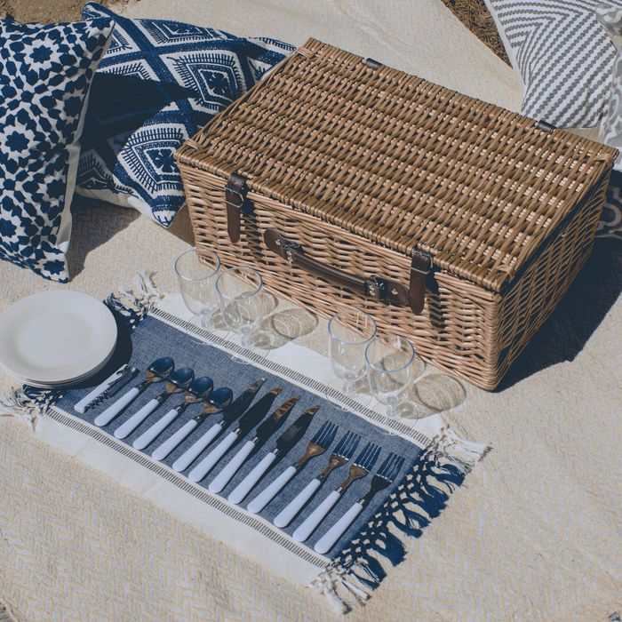 A wicker picnic basket placed on a blanket, surrounded by plates, glasses, and a complete cutlery set, all set for an outdoor feast