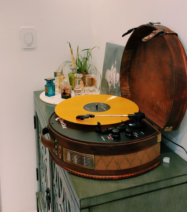 A reviewer’s image of a brown suitcase turntable with an orange vinyl spinning beneath a silver and black needle, resting on top of a green wooden cabinet with two black dials.