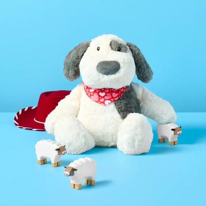 A plush dog toy adorned with a heart bandana, surrounded by tiny wooden sheep, set against a soft blue backdrop. A cowboy hat is subtly peeking out behind.