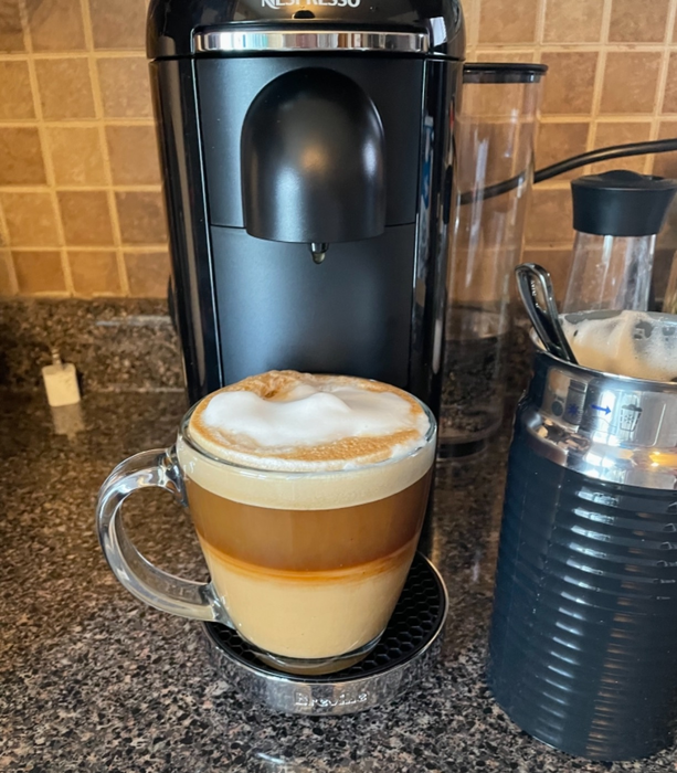 A glass mug filled with layered coffee and foam sits on a counter beside a coffee machine and milk frother.