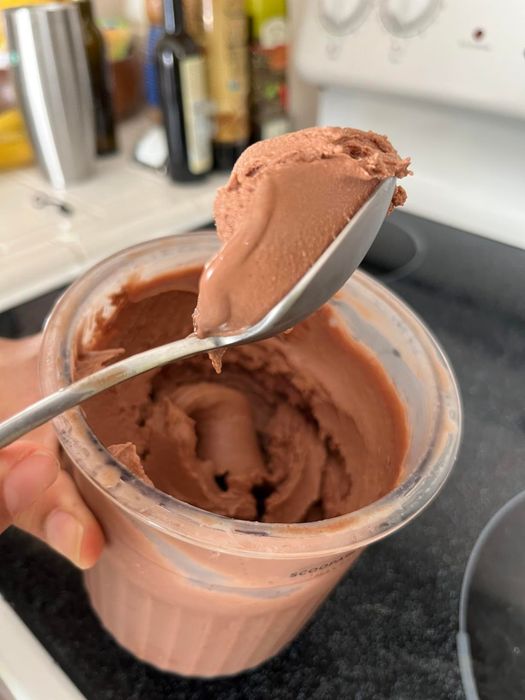 A spoon lifts smooth chocolate ice cream from a container placed on a kitchen counter, surrounded by various bottles and a stove.